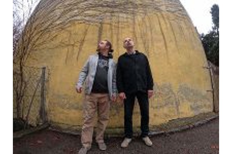 Das Bild zeigt zwei Männer, die vor einer gelben Wand stehen